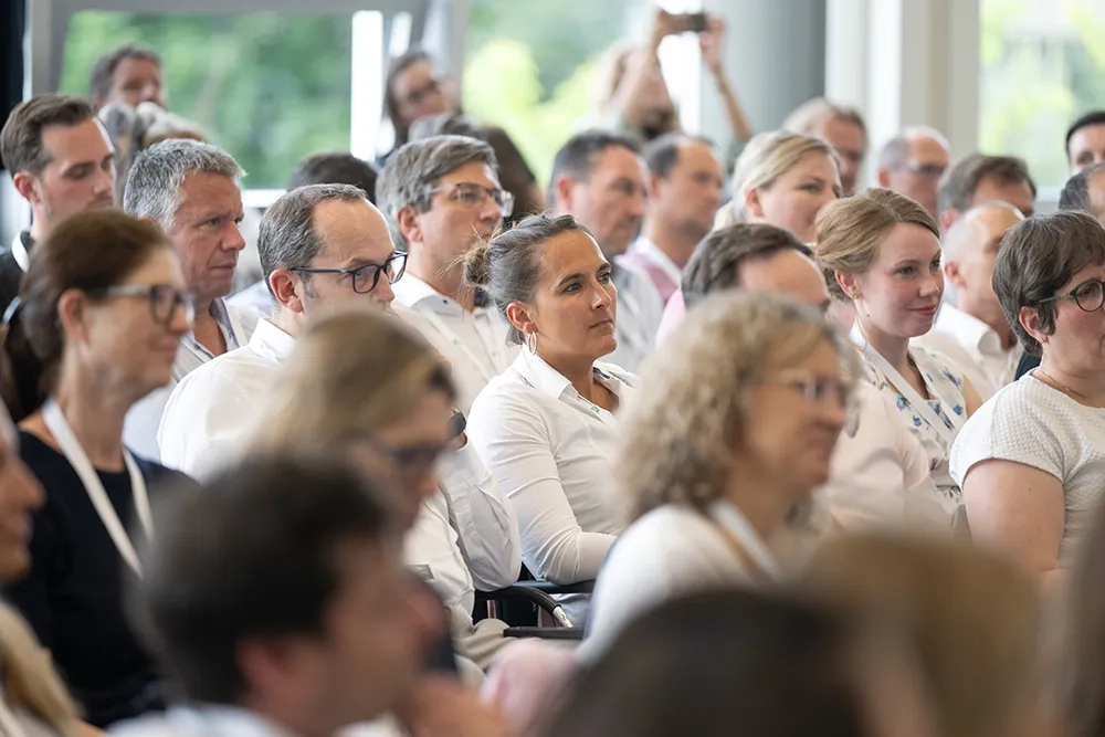 Ein dicht besetztes Publikum sitzt in den Reihen eines hellen Konferenzraums. Die Personen tragen überwiegend geschäftsmäßige Kleidung, wie helle Hemden und Blusen, und blicken aufmerksam in Richtung der Bühne. Im Hintergrund ist eine große Fensterfront mit Blick ins Grüne zu erkennen.