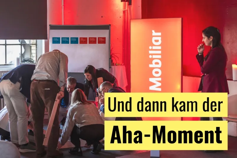 Redaktionelle Grafik mit dem Titeltext «Und dann kam der Aha-Moment» auf gelbem Balken im unteren Bildbereich: Eine Gruppe von Personen beugt sich bei einem Workshop konzentriert über ein gemeinsames Projekt auf dem Boden; im Hintergrund sind ein Flipchart mit farbigen Karten sowie ein beleuchtetes Mobiliar-Rollup in einem rot ausgeleuchteten Veranstaltungsraum zu sehen.