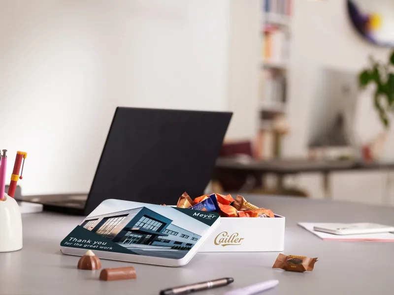 Eine geöffnete weiße Cailler-Pralinendose steht auf einem Schreibtisch. Der Deckel ist individuell bedruckt und zeigt ein modernes Firmengebäude, das Logo "Meyer" sowie den Text "Thank you for the great work". Die Dose ist mit einzeln verpackten Schokoladen gefüllt. Im unscharfen Hintergrund ist ein heller Büroarbeitsplatz mit einem Laptop zu erkennen.