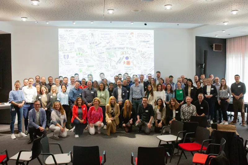 Gruppenfoto einer grossen Veranstaltung: Rund 80 Personen posieren in einem modernen Konferenzsaal vor einer grossformatigen Leinwand mit einem farbigen Graphic Recording zum Thema «From Constraint to Choice»; die Teilnehmenden tragen Namensschilder und sind in mehreren Reihen aufgestellt.