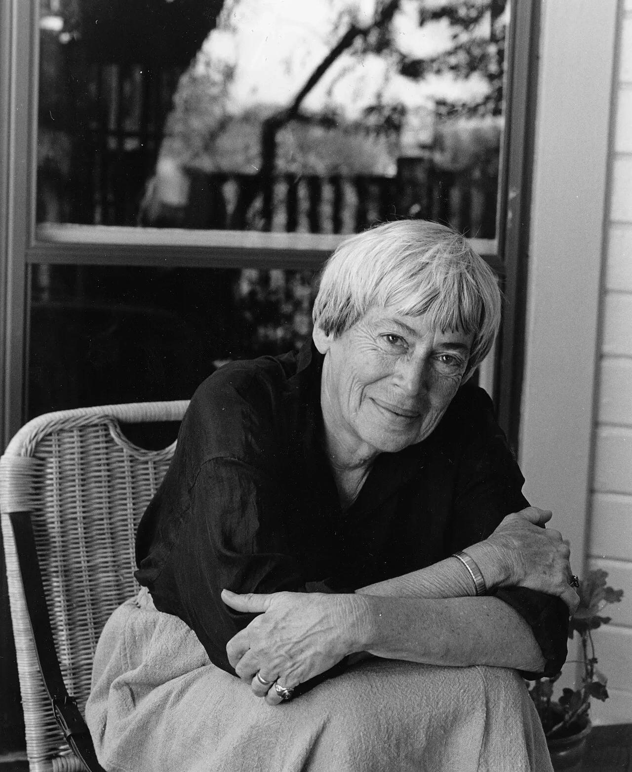 Eine Schwarz-Weiß-Fotografie der Autorin Ursula K. Le Guin. Sie sitzt entspannt in einem geflochtenen Korbstuhl, hat die Arme locker verschränkt und blickt mit einem sanften, herzlichen Lächeln direkt in die Kamera. Sie trägt eine dunkle Bluse und Ringe an den Fingern. Im Hintergrund ist ein großes Fenster zu sehen, in dessen Scheibe sich Bäume spiegeln.