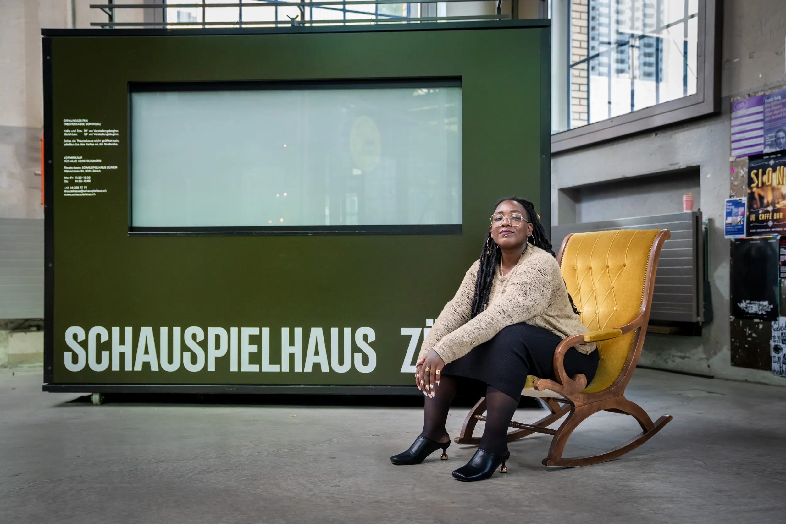 Yuvviki Dioh sitzt auf einem antiken Schaukelstuhl vor der Kasse des Schiffbaus. Sie sitzt selbstbewusst nach vorne gelehnt, trägt einen Strickpulli, einen schwarzen Rock und hohe Schuhe mit runden, goldenen Absätzen.