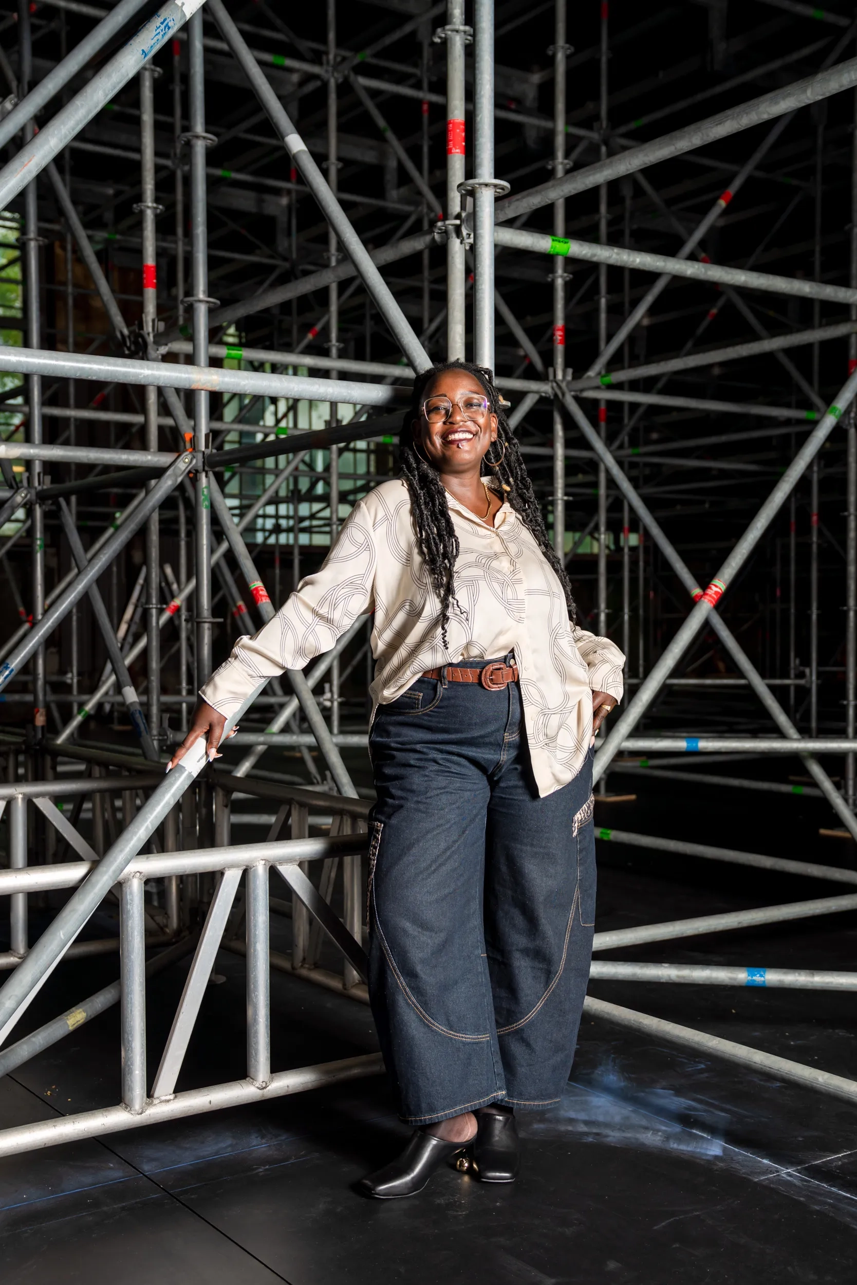 Yuvviki Dioh steht hinter der Schiffbau-Bühne, vor einem Gewirr aus Gerüsten, an die sie sich leicht anlehnt. Sie trägt blaue Jeans, einen braunen Gurt und eine hellbeige Bluse mit Knotenmuster. Sie lächelt breit in die Kamera.