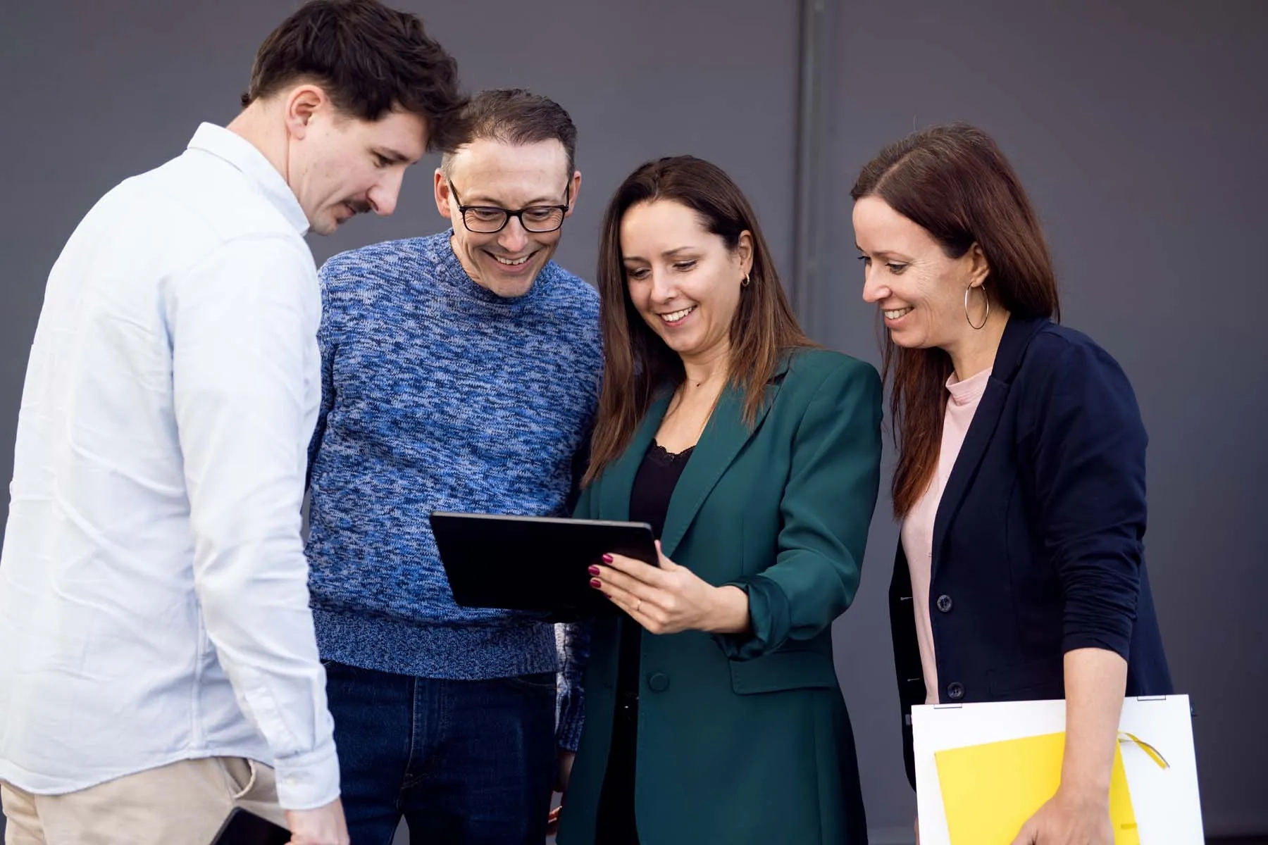 Eine Gruppe von vier Kolleginnen und Kollegen steht gut gelaunt vor einer neutralen, dunkelgrauen Wand. Eine Frau im Zentrum, die einen dunkelgrünen Blazer trägt, hält ein Tablet, auf das alle vier Personen gemeinsam blicken und dabei lächeln. Rechts im Bild hält eine weitere Frau in einem dunklen Sakko eine weiße Mappe, aus der ein gelbes Notizbuch herausschaut.