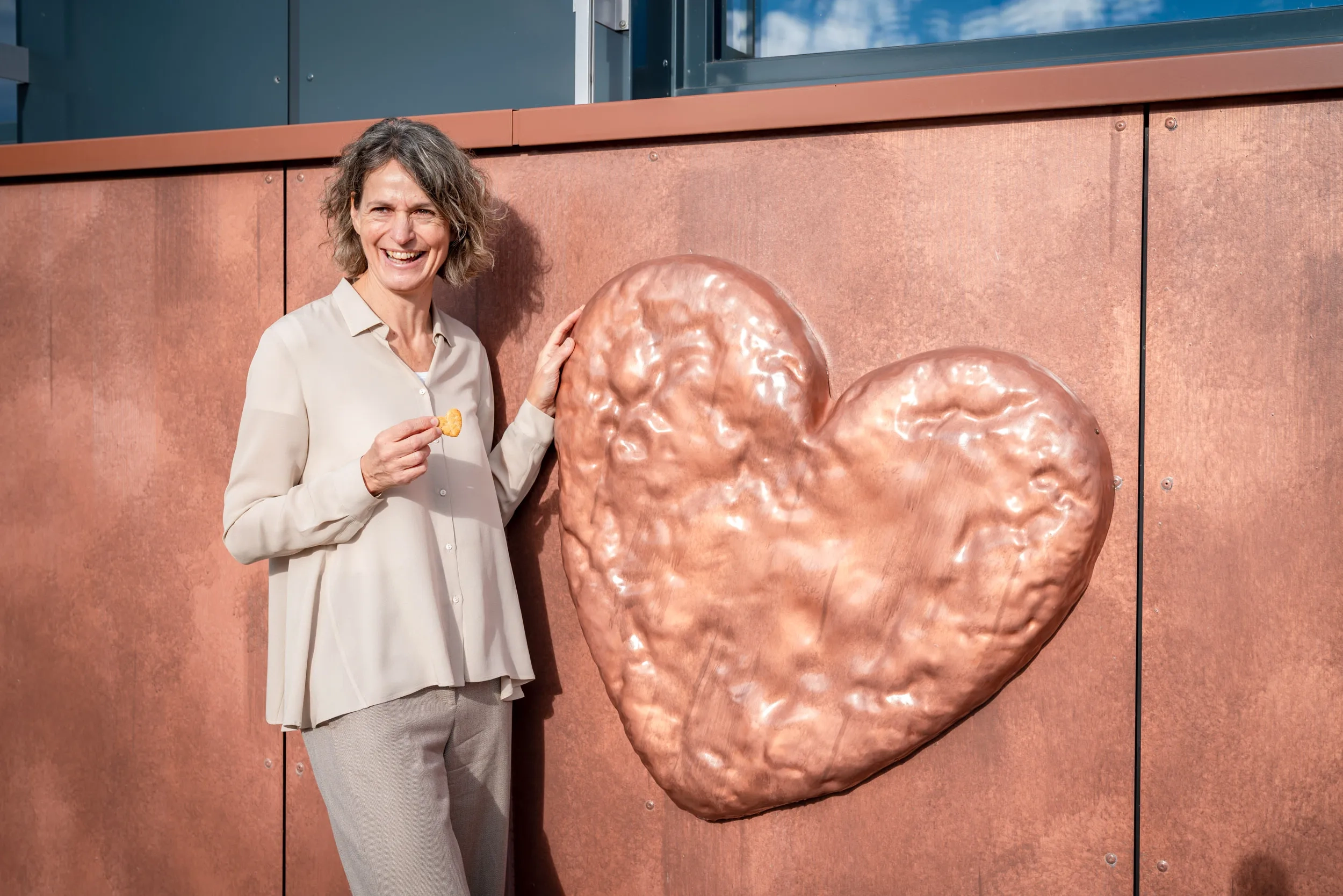 Yvonne Schorno steht lachend vor einer kupferfarbenen Wand, an der eine große, reliefartige Herz-Skulptur angebracht ist. Sie trägt eine helle Bluse und hält einen kleinen, herzförmigen Keks in der Hand, während sie ihre andere Hand leicht auf das große Metallherz legt.