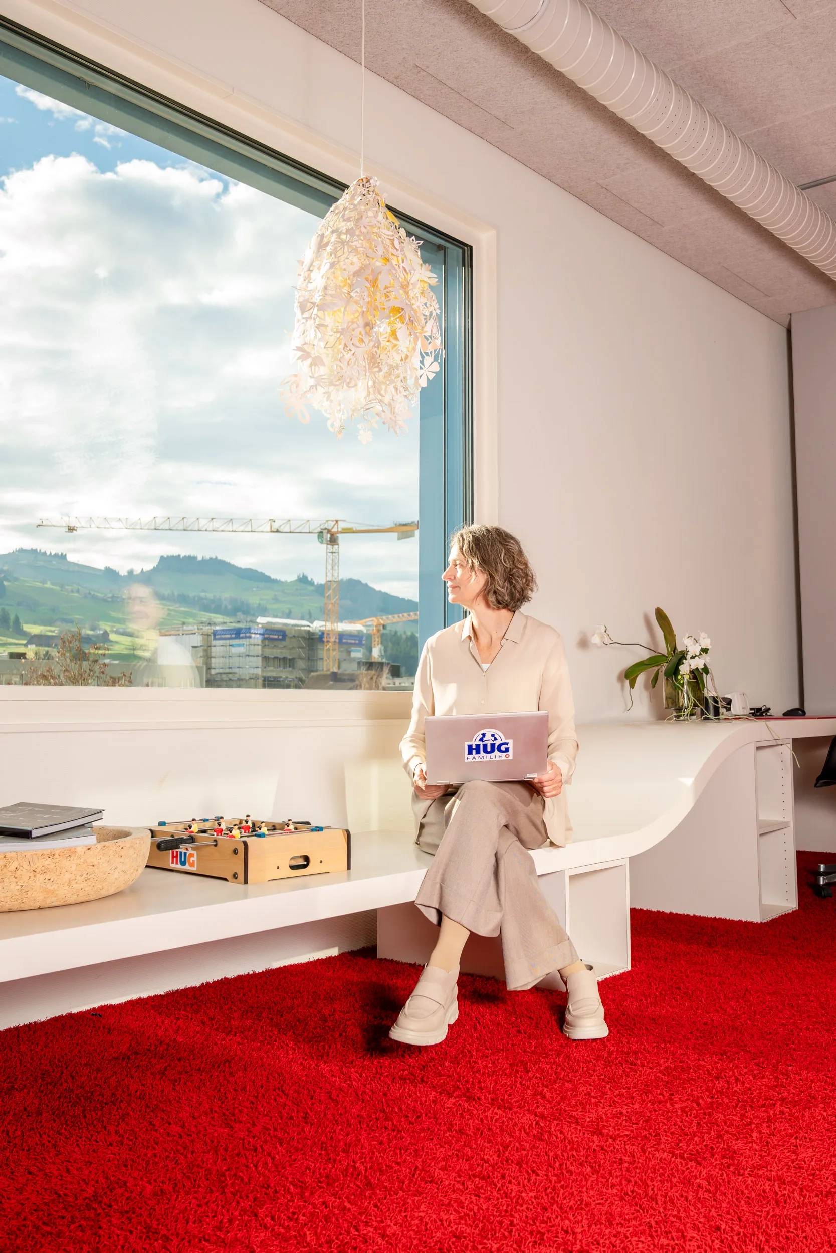 Yvonne Schorno sitzt lächelnd mit einem Laptop auf dem Schoß in einem modernen, hellen Büroraum auf einer geschwungenen weißen Sitzbank. Sie trägt helle Kleidung und blickt aus einem großen Fenster, das eine Aussicht auf eine hügelige Landschaft mit Baustellenkranen bietet. Der Raum ist mit einem auffälligen roten Teppich ausgelegt; auf der Bank neben ihr stehen ein kleiner Tischkicker und eine dekorative Schale. Auf dem Laptop ist ein Aufkleber der „HUG Familie“ sichtbar.