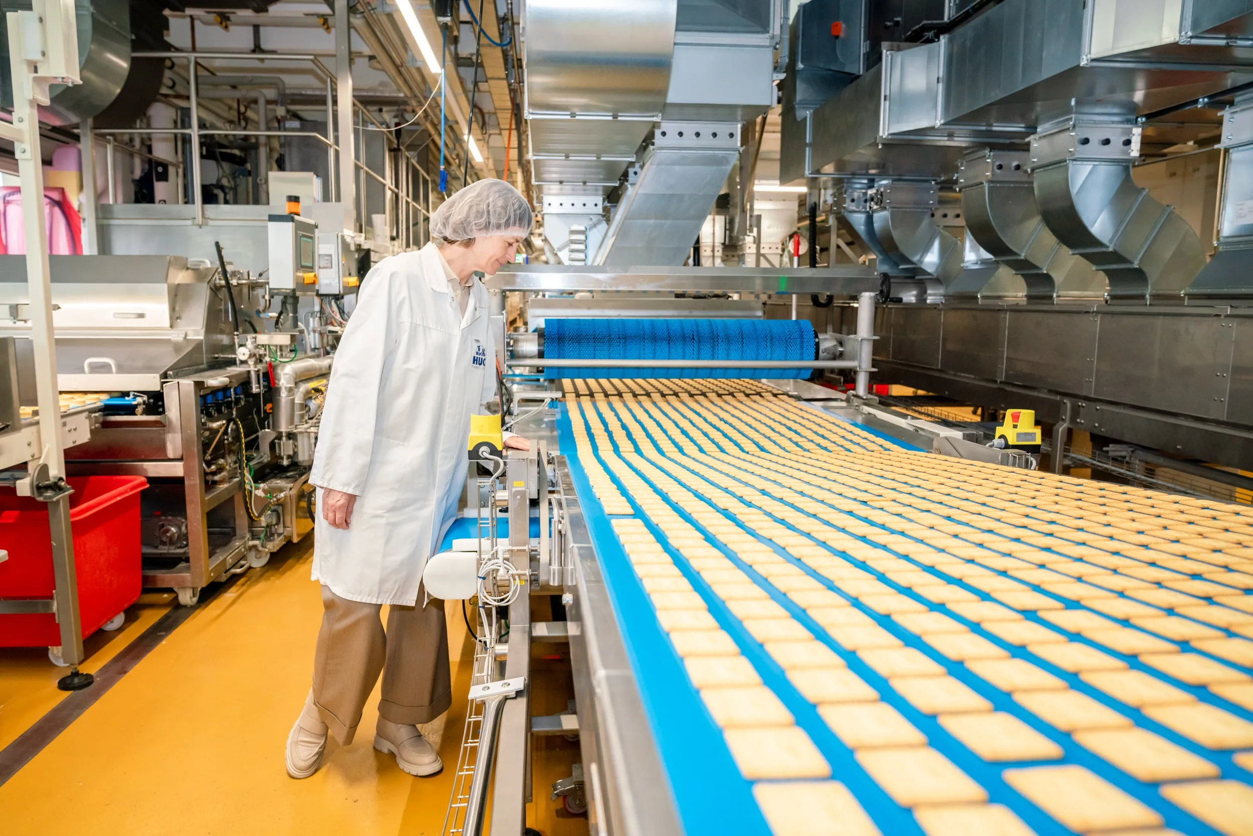 Yvonne Schorno steht in einer industriellen Produktionshalle an einem laufenden Förderband, auf dem hunderte kleiner, rechteckiger Kekse transportiert werden. Sie trägt vorschriftsgemäß einen weißen Arbeitsmantel und ein Haarnetz. Die Umgebung zeigt komplexe Maschinenanlagen und Rohrleitungen einer Backwarenfabrik.