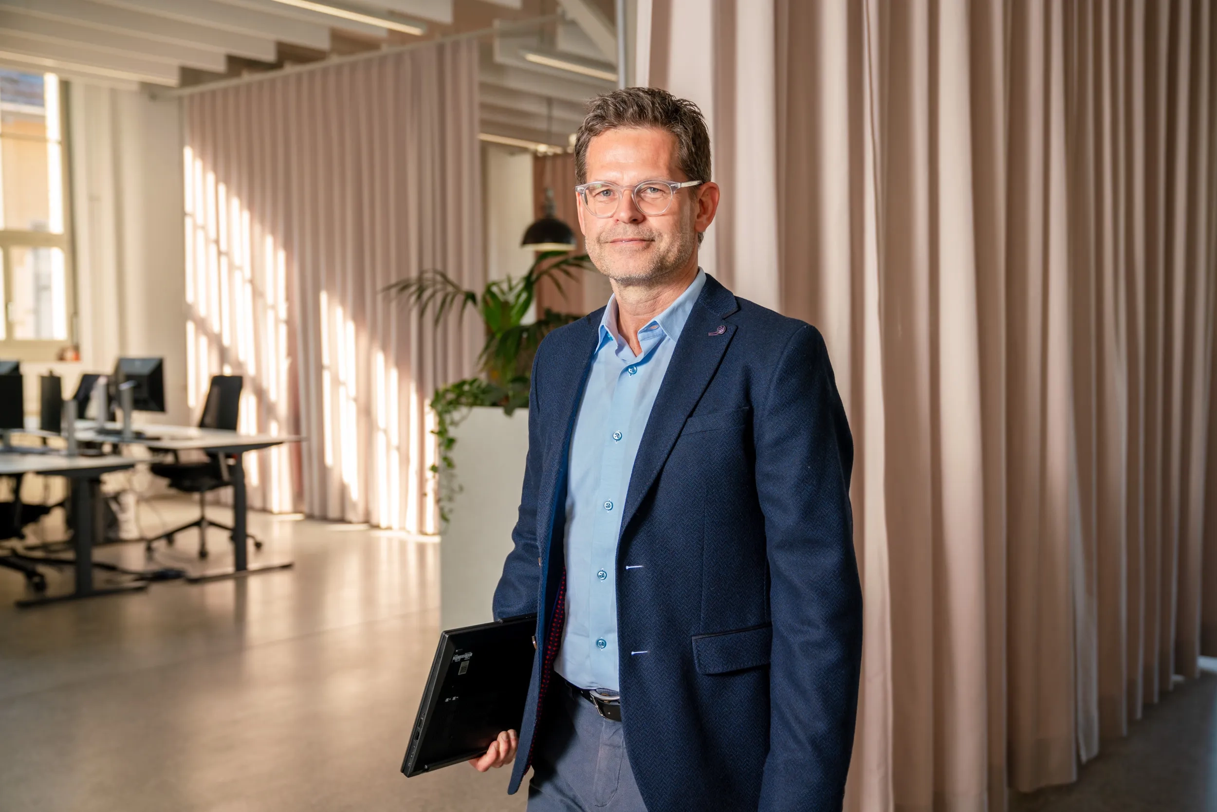 Thomas Biegel steht in einem modernen, hellen Büro. Er trägt eine Brille, ein hellblaues Hemd, ein dunkelblaues Sakko und eine graue Stoffhose. In seiner rechten Hand hält er einen geschlossenen, schwarzen Laptop. Er blickt mit einem leichten Lächeln direkt in die Kamera. Das Sonnenlicht fällt von links in den Raum. Im unscharfen Hintergrund sind auf der linken Seite leere Schreibtische mit Monitoren zu erkennen, in der Mitte steht eine grüne Zimmerpflanze und auf der rechten Seite hängen bodenlange, beigefarbene Vorhänge.