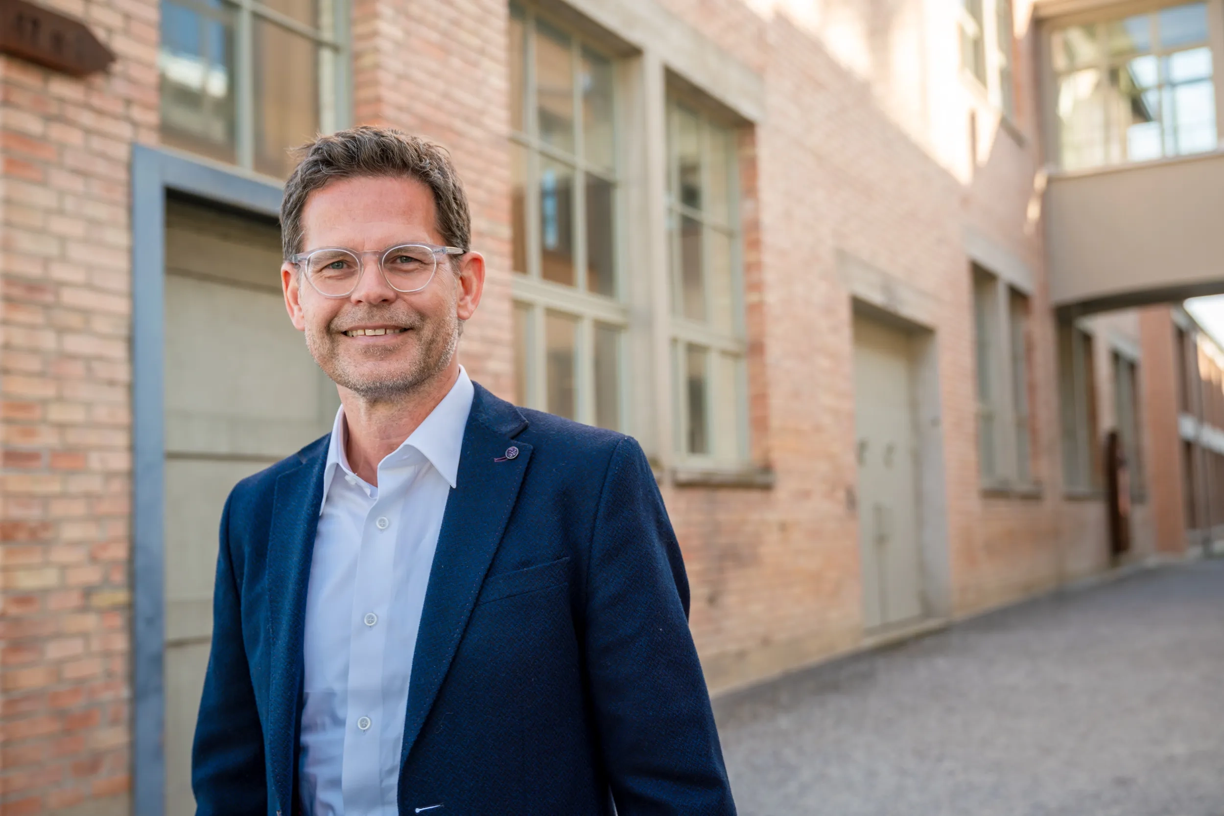 Porträt von Thomas Biegel. Er trägt eine Brille, ein weißes Hemd sowie ein dunkelblaues Sakko und blickt freundlich lächelnd in die Kamera. Im unscharfen Hintergrund ist ein helles Backsteingebäude zu sehen.