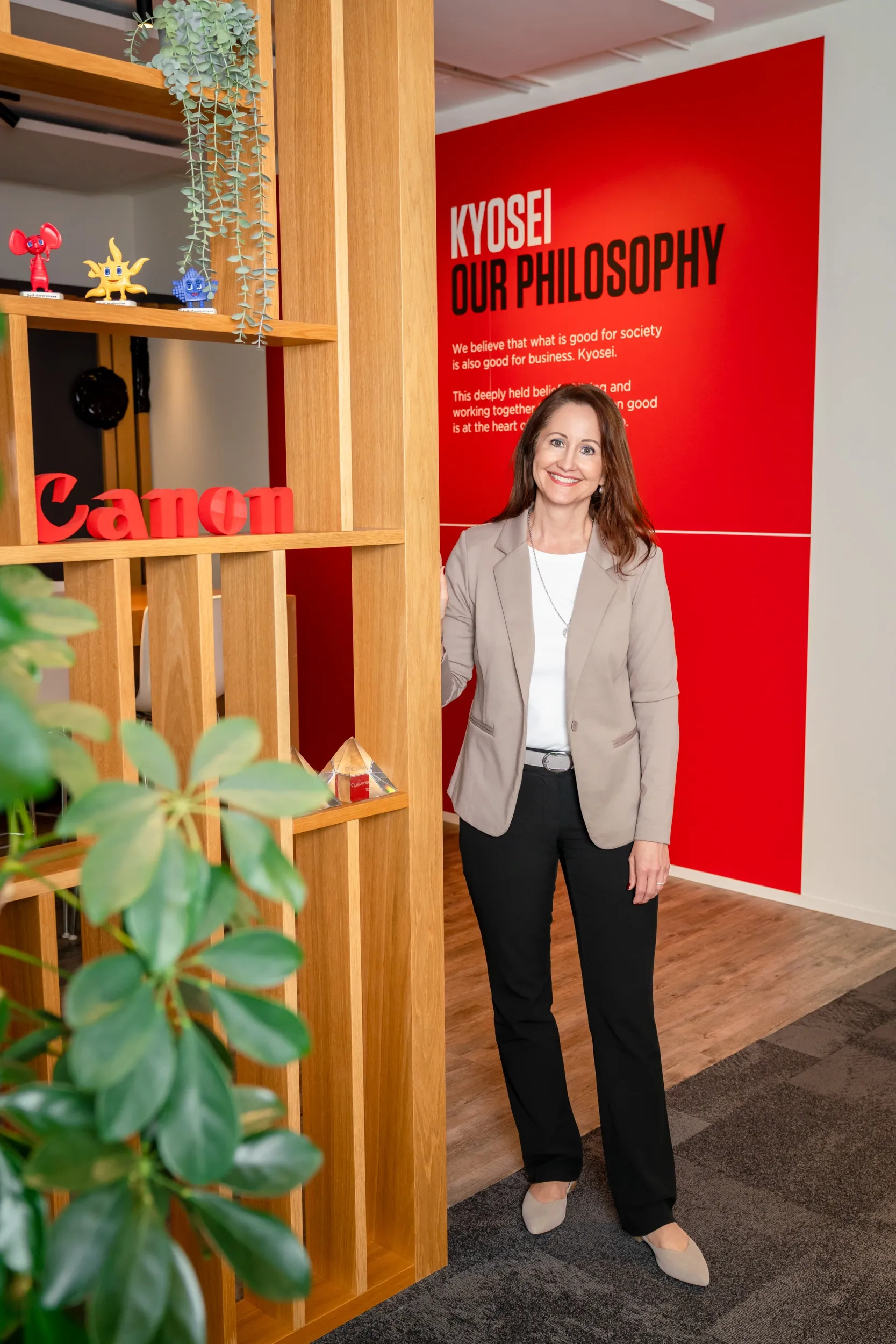 Corinne von Felten steht lächelnd im Eingangsbereich der Canon-Schweiz-Niederlassung neben einem Holzregal mit dem roten Canon-Schriftzug und kleinen Figuren; im Hintergrund eine rote Wand mit dem Text «Kyosei — Our Philosophy».