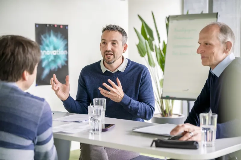 Thomas Sepassi (Mitte) und Christoph Widmer (rechts) im Gespräch mit einer dritten Person am Besprechungstisch; Sepassi spricht mit lebhafter Gestik, auf dem Tisch stehen Wassergläser und Unterlagen, im Hintergrund sind ein Flipchart und ein «Innovation»-Poster zu sehen.