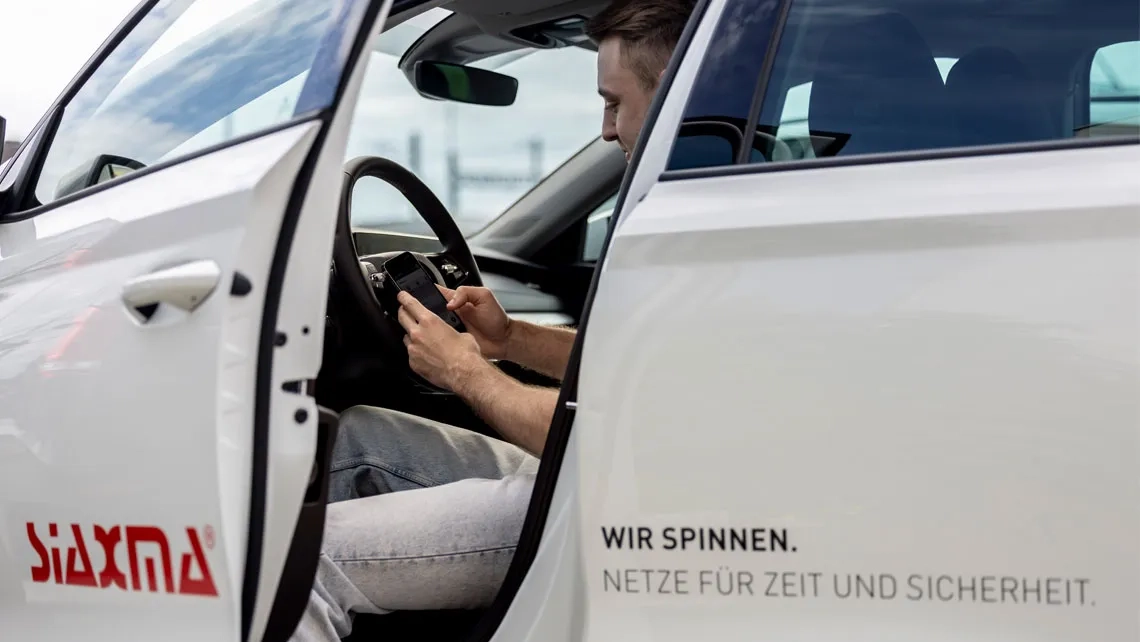 Ein junger Mann sitzt auf dem Fahrersitz eines weißen Autos, die Fahrertür steht weit offen. Er blickt konzentriert auf sein Smartphone, das er mit beiden Händen hält. Auf der weißen Karosserie des Autos ist links das rote Logo der Firma „SIAXMA“ zu sehen. Auf der Tür steht der Slogan: „WIR SPINNEN. NETZE FÜR ZEIT UND SICHERHEIT.“ Der Hintergrund zeigt einen leicht bewölkten Himmel.