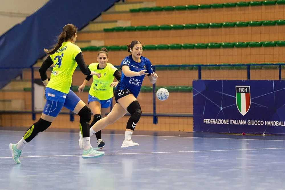 Handballspielszene in einer Sporthalle: Eine Spielerin im blauen Trikot mit der Aufschrift „ITALIA HANDBALL“ läuft mit dem Ball in Richtung Tor, verfolgt von zwei Gegnerinnen im gelb-blauen Trikot. Im Hintergrund sind leere grüne Tribünensitze und eine Werbebande mit dem Logo und Schriftzug der „Federazione Italiana Giuoco Handball“ (FIGH) zu sehen.