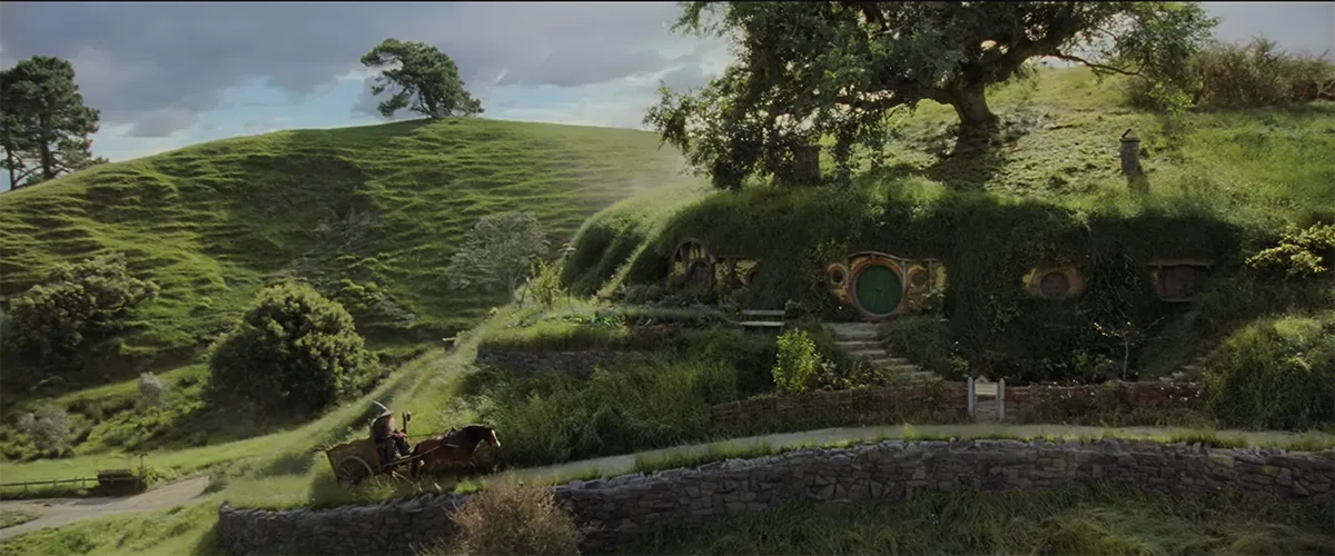 Eine friedliche, sonnendurchflutete Landschaftsaufnahme aus dem Auenland im ersten „Herr der Ringe“-Film. Im Zentrum und auf der rechten Seite sieht man die Hobbit-Höhle Beutelsend, die direkt in einen saftig grünen, grasbewachsenen Hügel gebaut ist. Sie hat eine markante runde, grüne Tür, kleine runde Fenster und einen großen, alten Baum mit üppiger Krone, der direkt oben auf dem Dach des Hügels wächst. Vor der Tür führt eine Steintreppe zu einem geschwungenen Weg hinab, der von niedrigen Steinmauern und Holzzäunen gesäumt ist. Auf diesem Weg links unten nähert sich Gandalf der Graue, erkennbar an seinem grauen Spitzhut. Er sitzt auf einem kleinen Holzkarren, der von einem braunen Pferd den Hügel hinaufgezogen wird. Die gesamte Szenerie mit ihren sanften, grünen Hügeln im Hintergrund strahlt eine tiefe, ländliche Idylle aus.