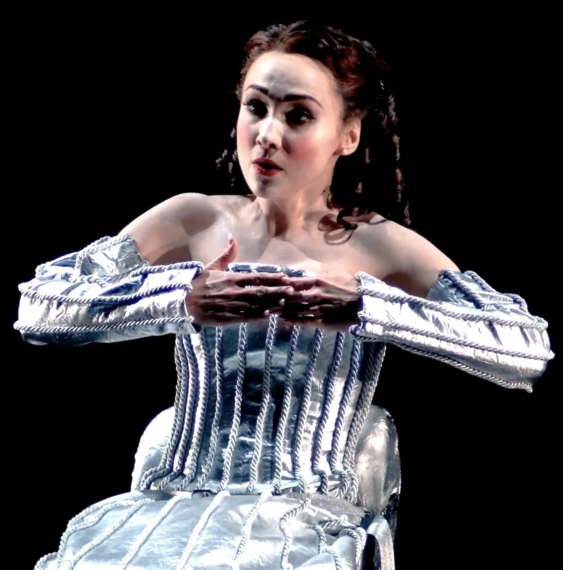 Die Opernsängerin Cristina Păsăroiu in der Rolle der Olympia an der Deutschen Oper Berlin (2018). Sie ist vor einem komplett schwarzen Hintergrund zu sehen und trägt ein futuristisches, silbernes Kostüm. Es besteht aus einem starren, metallisch wirkenden Korsett und separaten Ärmeln, die mit silbernen Kordeln verziert sind. Ihre Haltung ist roboterhaft und steif, die Hände sind vor der Brust zusammengeführt. Sie ist blass geschminkt und blickt mit einem puppenhaften, leeren Ausdruck leicht nach oben.