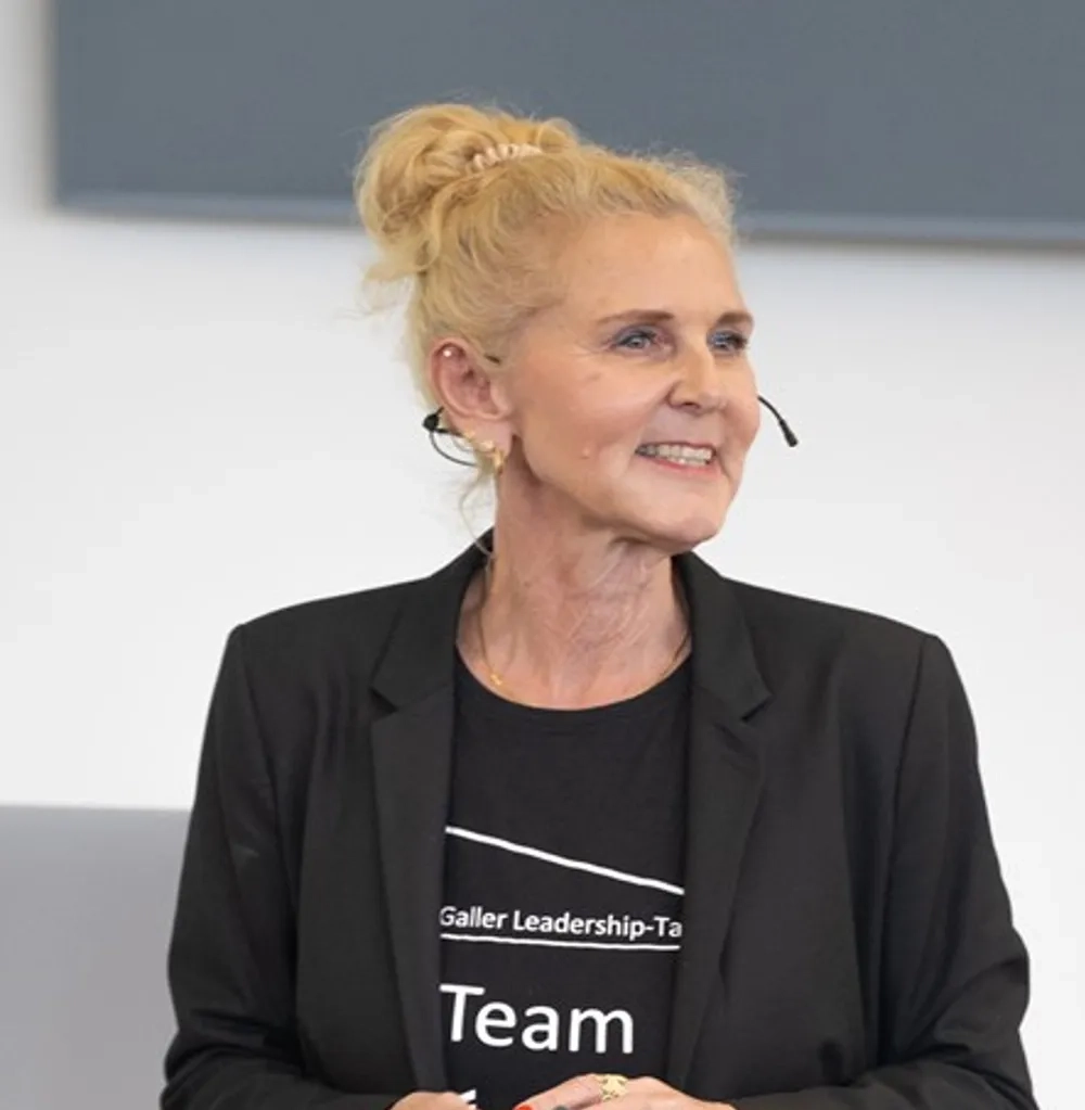 Porträtaufnahme einer lächelnden Frau (Heike Bruch) mit blonden, zu einem Dutt hochgesteckten Haaren. Sie trägt ein unauffälliges Headset-Mikrofon sowie einen schwarzen Blazer über einem schwarzen T-Shirt mit der Aufschrift „Team“ und blickt aufmerksam zur Seite.