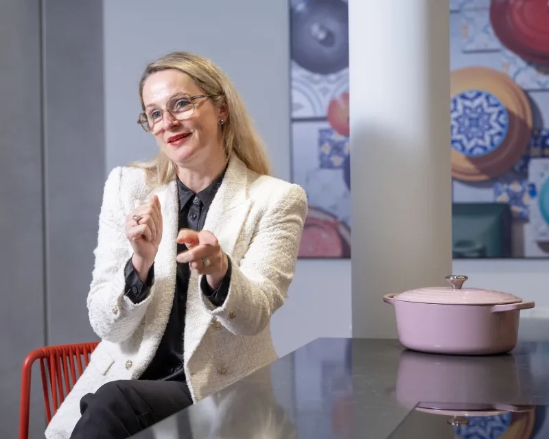 Nahaufnahme von Maryline Tissot, die mit Brille und rotem Lippenstift lächelnd an einer dunklen Arbeitsplatte sitzt und mit den Händen gestikuliert. Rechts neben ihr steht ein rosa Le-Creuset-Bräter auf dem Tisch.