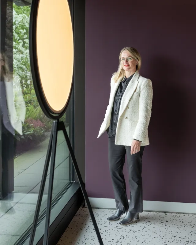 Ganzkörperporträt von Maryline Tissot in weißem Tweed-Blazer, schwarzer Hose und Ankle Boots. Sie steht neben einer großen Ringleuchte auf einem Stativ vor einer dunkelvioletten Wand auf Terrazzoboden.