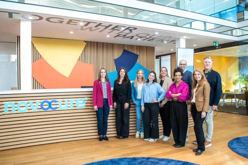 Eine Gruppe von neun lächelnden Personen steht für ein Gruppenfoto in einer hellen, modernen Büro-Lobby. Sie posieren vor einem Empfangstresen mit Holzlamellen und dem hellblauen Firmenlogo "novocure". An der Deckenstruktur über ihnen ist der Schriftzug "TOGETHER WITH OUR PATIENT" teilweise zu lesen. Die Personen sind unterschiedlich gekleidet, überwiegend im Business-Casual-Stil.