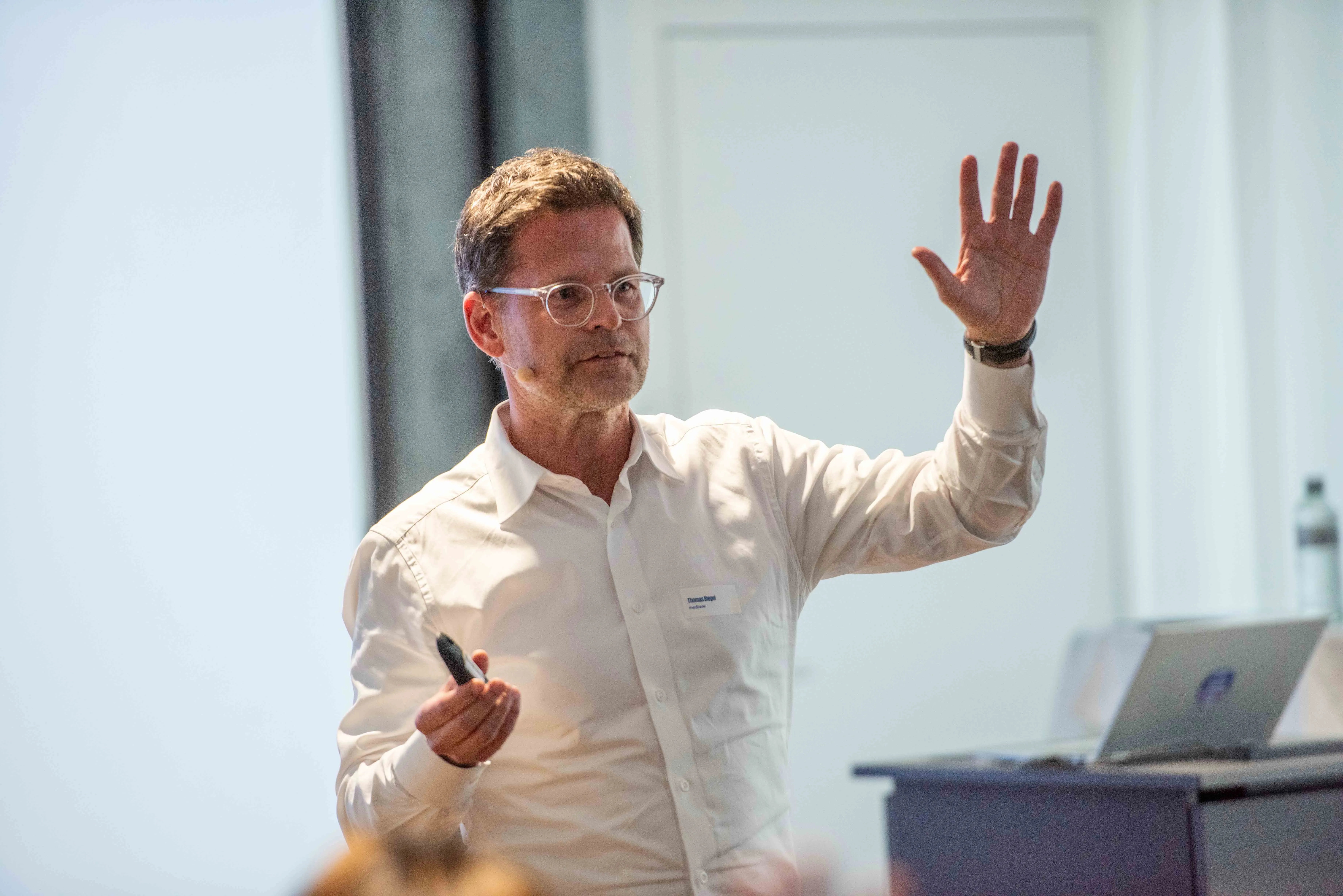 Thomas Biegel, ein Mann mit Brille in einem weißen Hemd spricht während eines Vortrags. Er hält einen Presenter in der rechten Hand und hat die linke Hand erklärend erhoben. Er trägt ein dezentes Headset-Mikrofon; im Hintergrund ist ein Laptop auf einem Pult erkennbar.