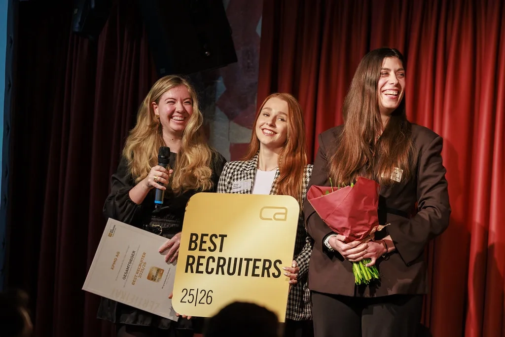 Drei freudestrahlende Frauen stehen bei einer Preisverleihung auf einer Bühne vor einem roten Vorhang. Sie halten ein Mikrofon, eine Urkunde für die KPMG AG als Gesamtsieger, ein großes goldenes Schild mit der Aufschrift 'BEST RECRUITERS 25|26' und einen roten Blumenstrauß.
