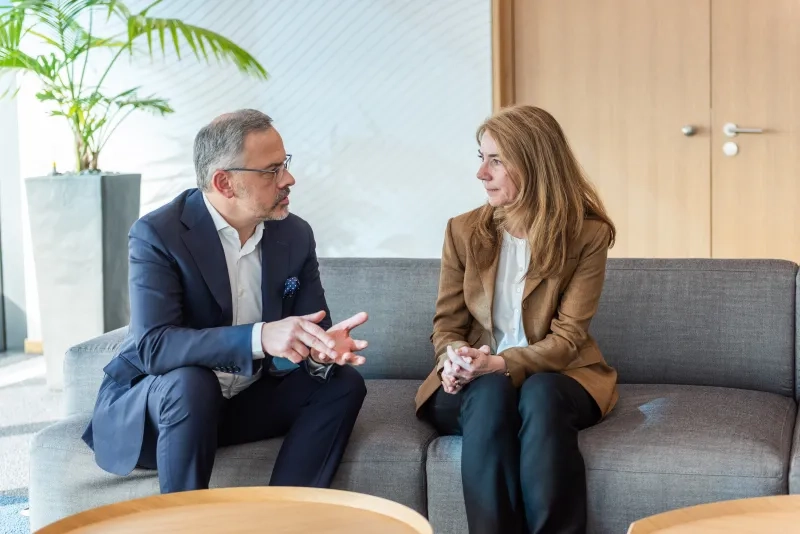 Michael Puri, ein Mann im dunkelblauen Anzug und eine Frau im braunen Blazer sitzen auf einem grauen Sofa in einem hellen Raum und führen ein Gespräch. Der Mann spricht und gestikuliert mit den Händen, während die Frau, Nancy Brunner, ihm aufmerksam zuhört.