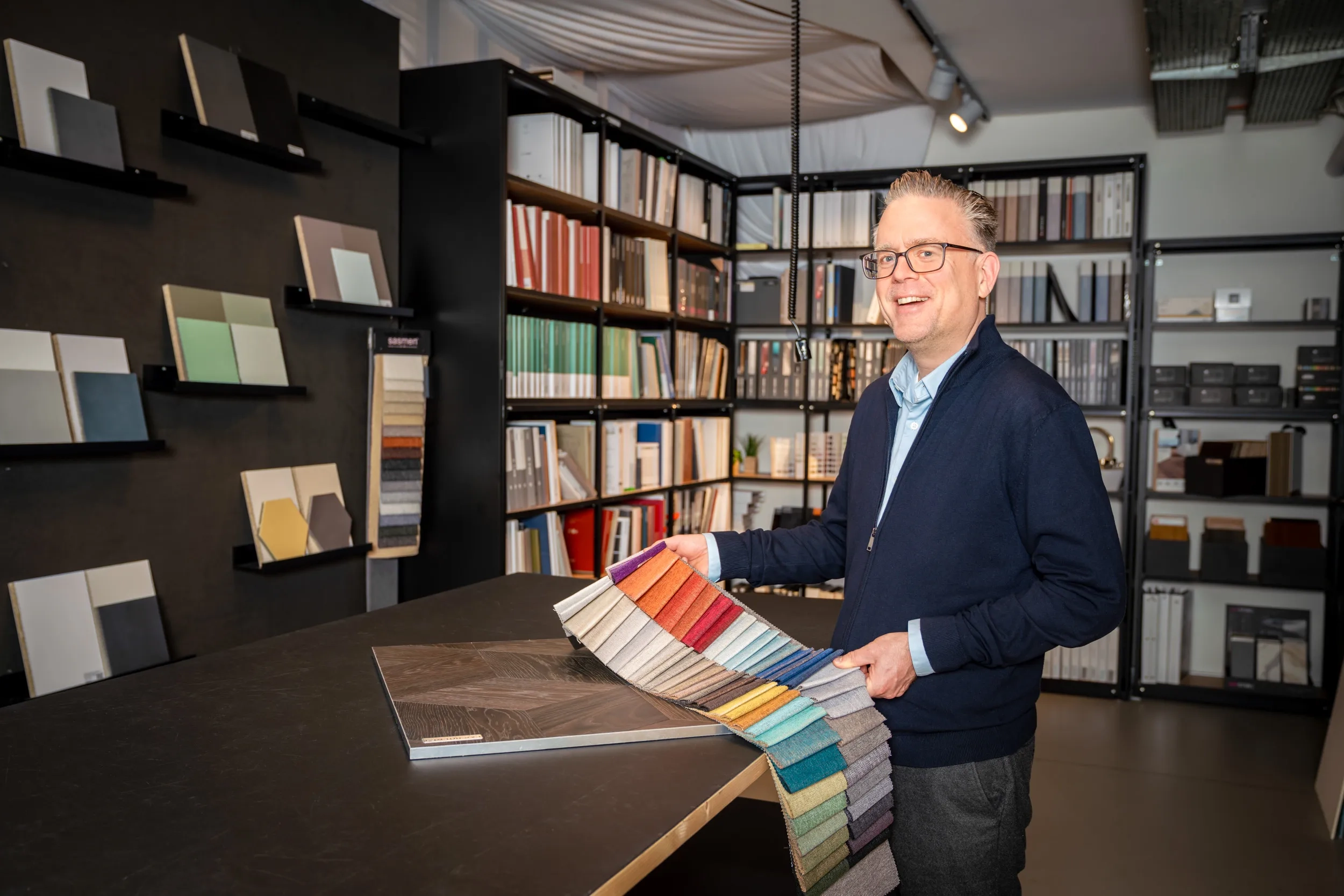 Ein Mann mit Brille und dunkelblauer Strickjacke steht lächelnd in einer modern eingerichteten Materialbibliothek. Er hält eine grosse Farbmusterpalette mit verschiedenen Stoffen in der Hand, die auf einem Tisch ausgebreitet ist. Im Hintergrund befinden sich Regale mit zahlreichen Musterbüchern, Ordnern und Materialproben, darunter Holz-, Farb- und Textilmuster.