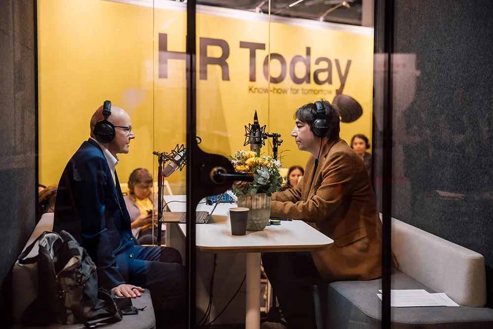 Daniel Thüler, Chefredaktor von HR Today (rechts), und Christian Hunziker von SwissICT (links) sitzen sich in einer schallgedämmten Aufnahmekabine gegenüber und sprechen in Mikrofone. Beide tragen Kopfhörer. Im Hintergrund ist das gelbe HR Today Logo zu sehen, und durch das Glas sind einige Zuschauer:innen erkennbar. Auf dem Tisch stehen Blumen, Notizen und Kaffeebecher.