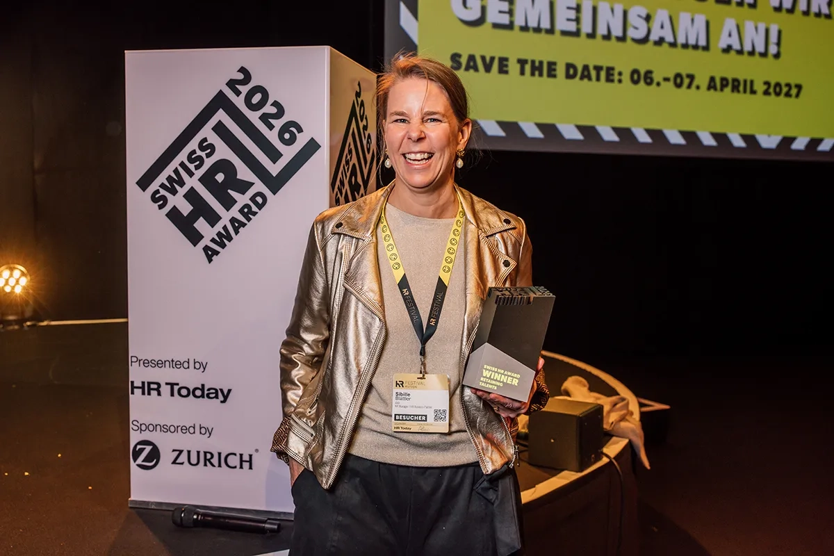 Eine lachende Frau in einer glänzenden Metallic-Jacke und mit einem Event-Lanyard um den Hals steht auf der Bühne. Sie hält eine schwarze Trophäe mit der Aufschrift "Swiss HR Award Winner Retaining Talents" in der Hand.