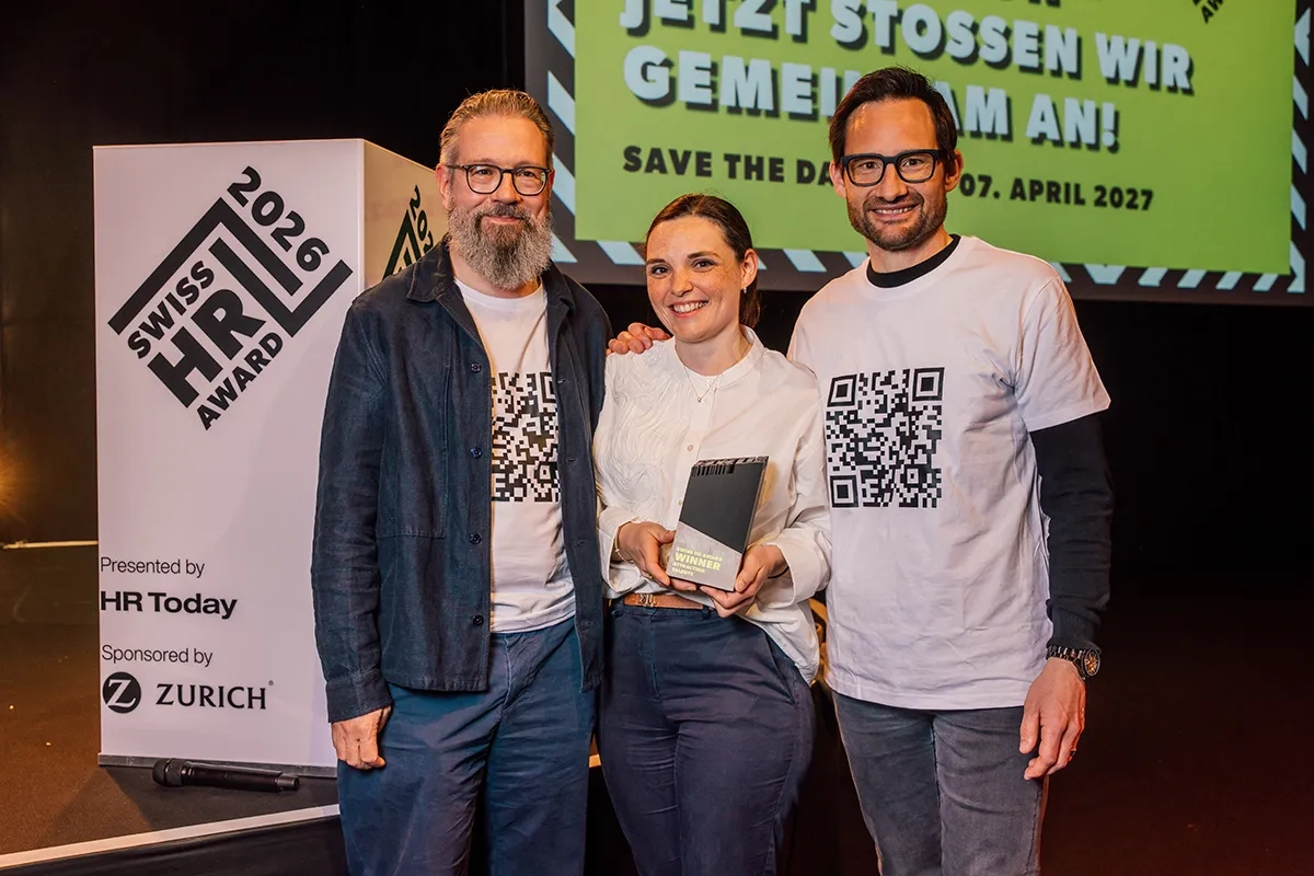 Drei Personen stehen bei der Verleihung des Swiss HR Award 2026 auf der Bühne. Eine Frau in der Mitte hält lächelnd die Trophäe, flankiert von zwei Männern, die weiße T-Shirts mit großen, aufgedruckten QR-Codes tragen.