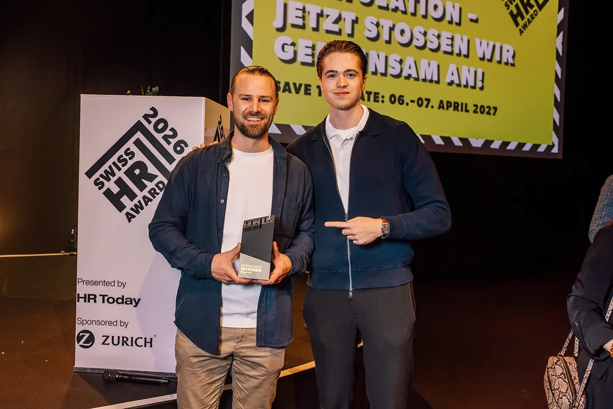 Zwei Männer stehen nach einer Preisverleihung auf der Bühne. Der linke Mann hält eine schwarze Trophäe ("Swiss HR Award Winner Start-Up") in den Händen, während der rechte Mann lächelnd auf ihn und den Preis zeigt.