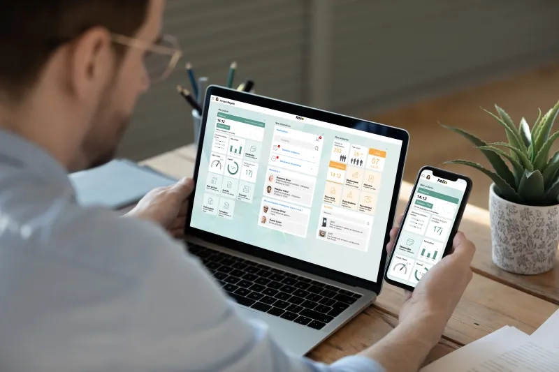 Eine Person von hinten betrachtet arbeitet gleichzeitig an einem Laptop und hält ein Smartphone in der Hand; auf beiden Bildschirmen ist eine HR-Software-Oberfläche mit Dashboards, Mitarbeiterprofilen und Diagrammen zu sehen.